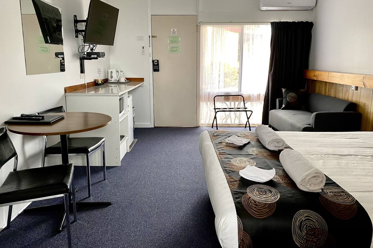 Ein Hotelzimmer mit einem Bett, einem Schreibtisch mit Stuhl, einer kleinen Küchenzeile, einem Fernseher und einem Fenster mit Vorhängen.