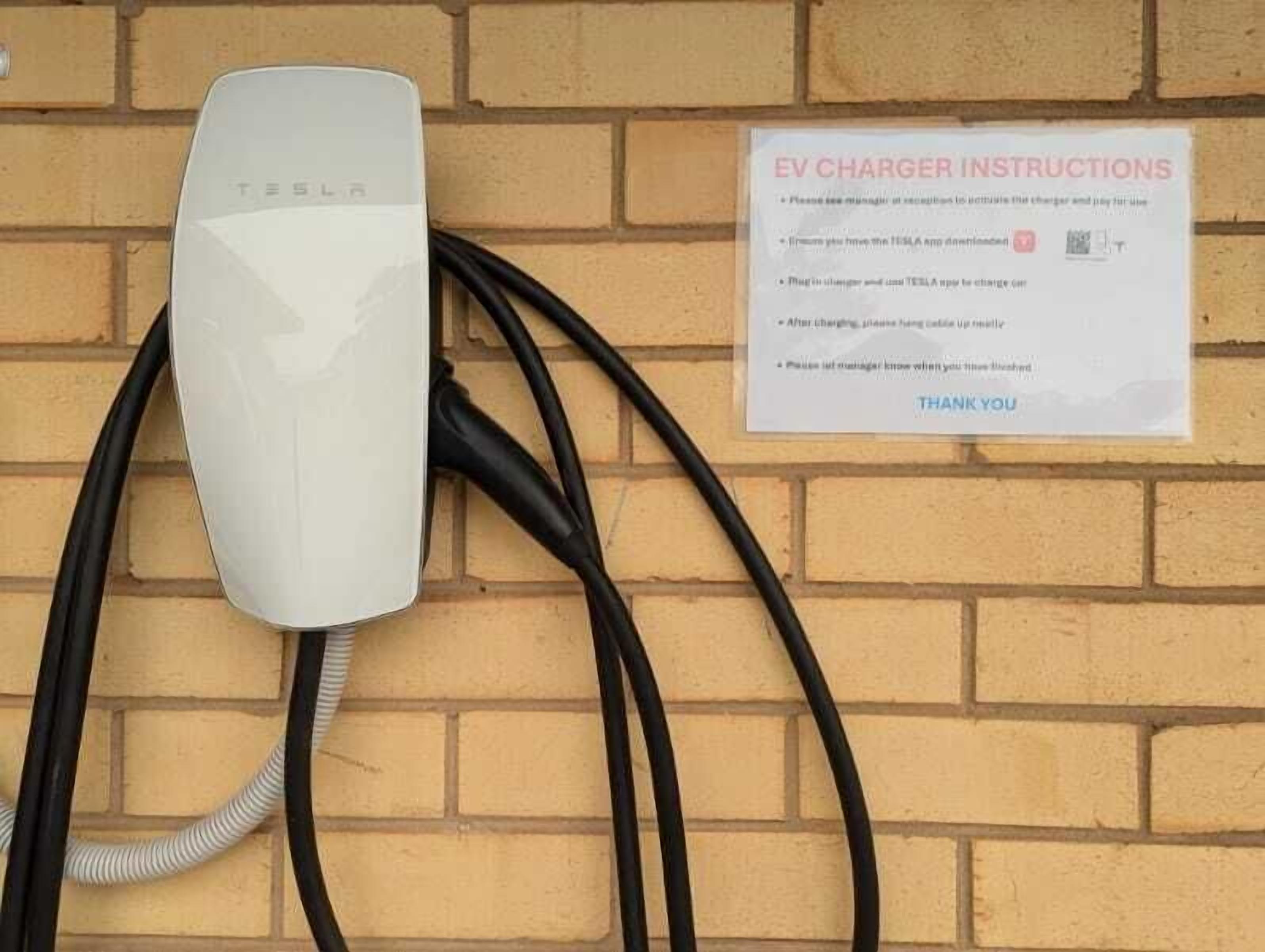 Une borne de recharge pour véhicules électriques Tesla est fixée à un mur en briques, et une pancarte à proximité indique les instructions à suivre.