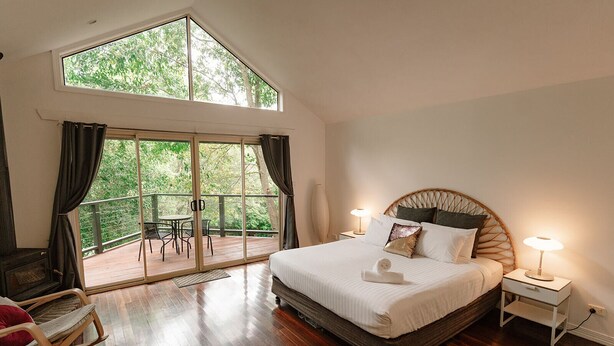 Un dormitorio con una cama grande, una mesita de noche, una lámpara, piso de madera, un balcón con mesa y sillas, y vistas a zonas verdes.
