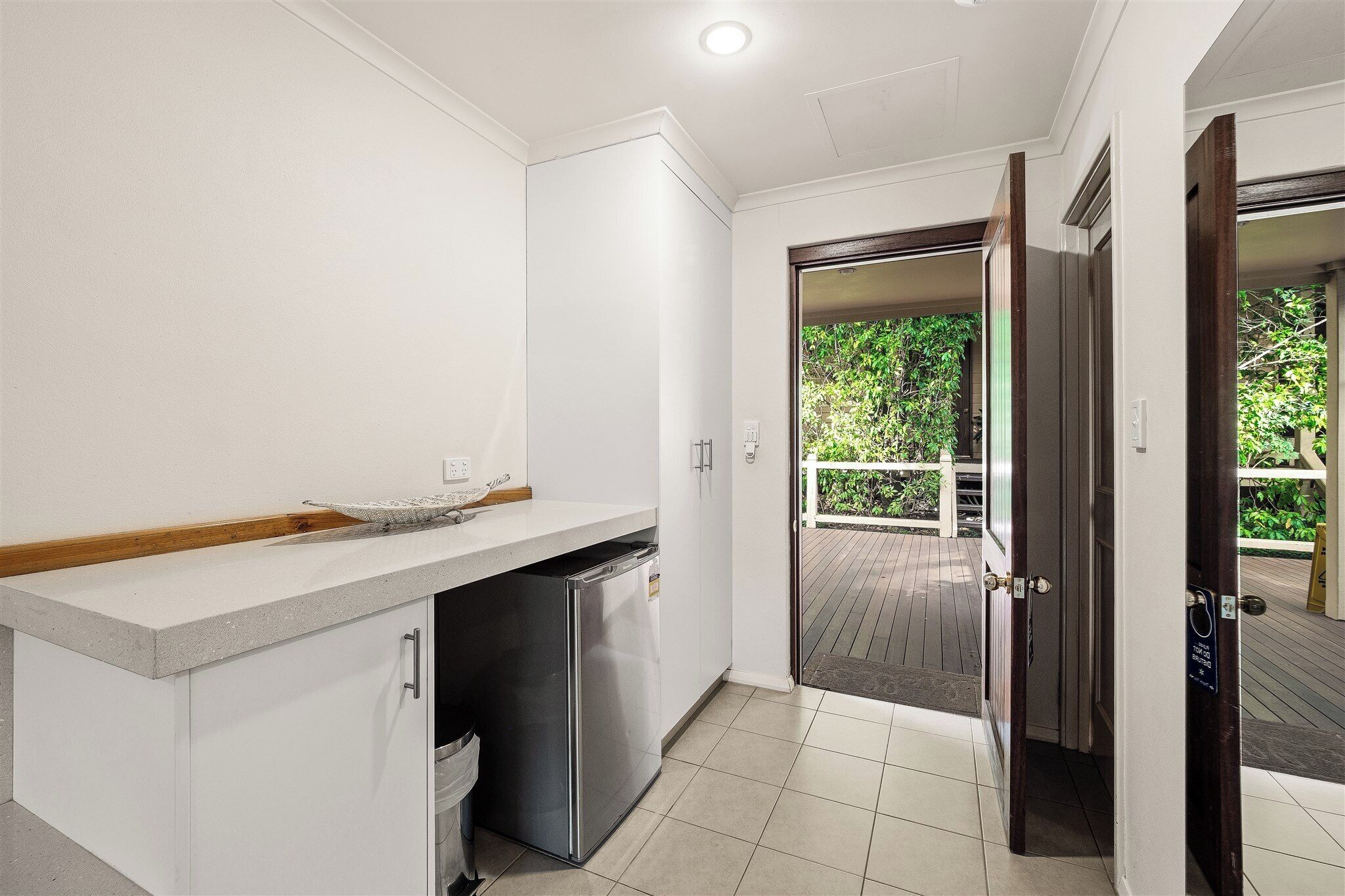 Une cuisine compacte équipée d’un réfrigérateur intégré, d’un petit évier et d’un plan de travail. Une porte donne accès à un espace extérieur doté d’une terrasse en bois.