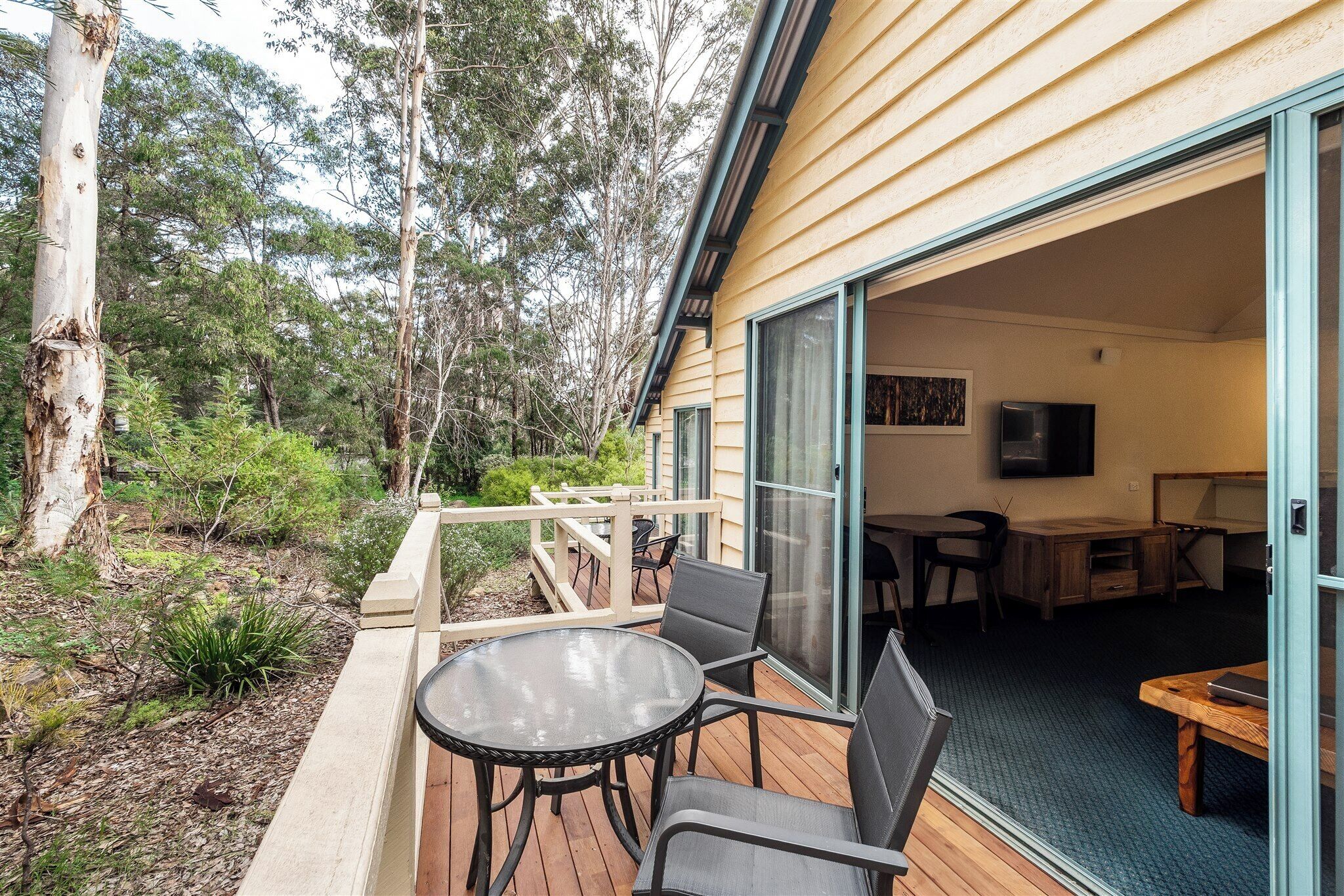 Una terraza de madera con un ventanal de vidrio que da a una habitación con escritorio, silla y televisor.