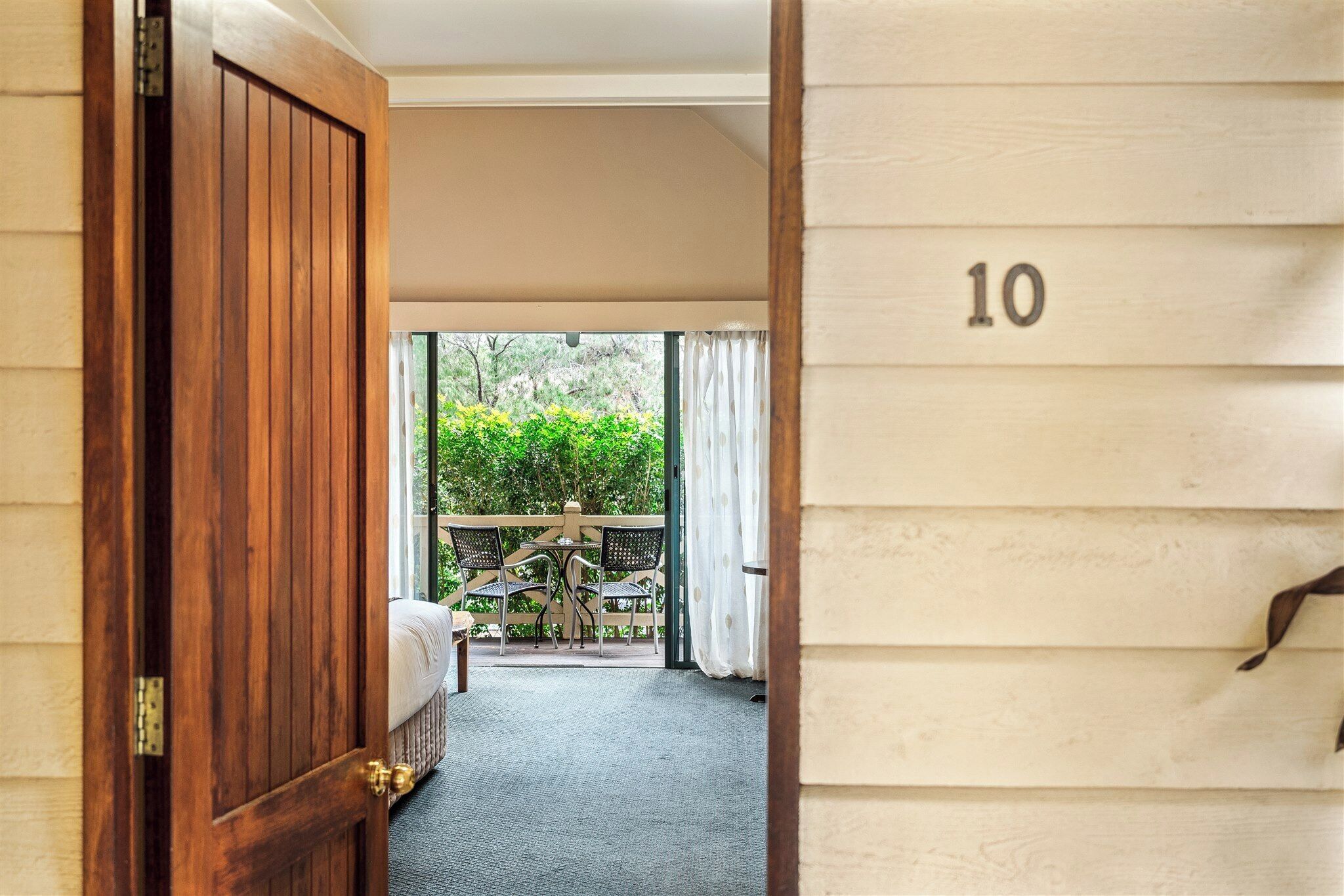 A hotel room with a wooden door, a number '10' on the wall, a bed, a small table, and a view of greenery outside.