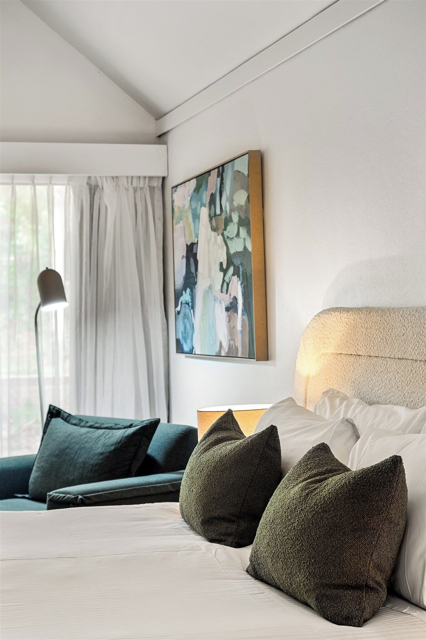 A modern hotel room with a bed, green pillows, a painting, and a lamp.