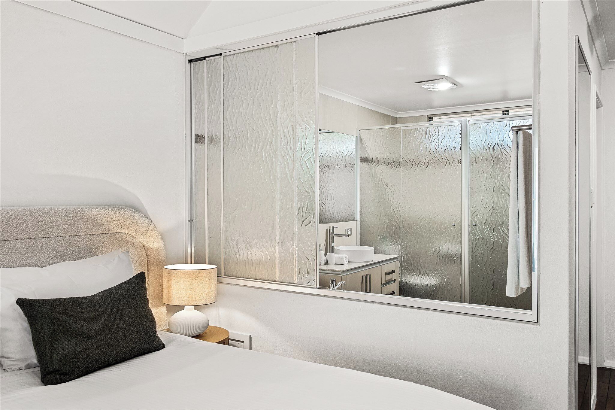 Ein Hotelzimmer mit Bett, Nachttisch, Lampe, großem Spiegel und einem Badezimmer mit Dusche und Waschbecken.