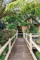 A wooden bridge leading to a building with a traditional design, surrounded by lush greenery and a car parked nearby.