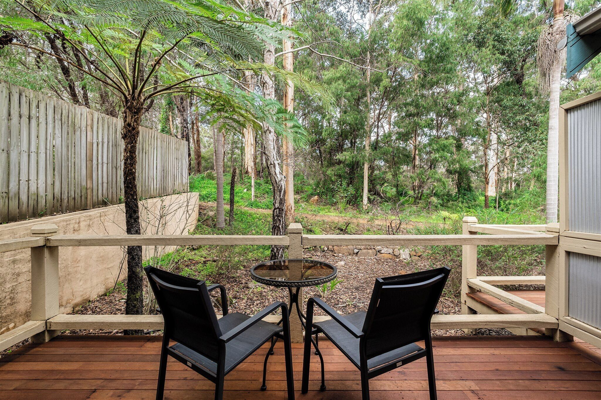 Eine Holzterrasse mit zwei Stühlen und einem Tisch, mit Blick auf ein bewaldetes Gebiet mit Holzzaun und einem Gebäude im Hintergrund.