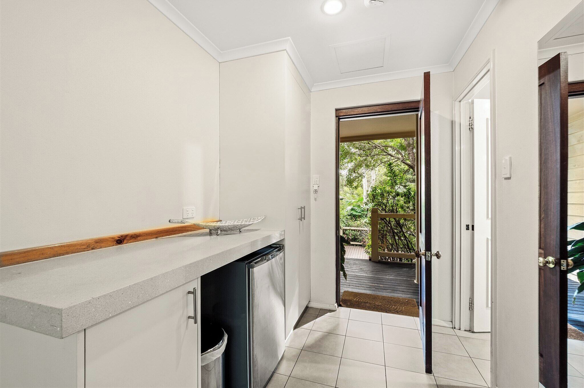 A narrow hallway with a kitchen area on the left, featuring a countertop and a dishwasher. A door on the right leads to an outdoor area with a wooden deck and greenery.