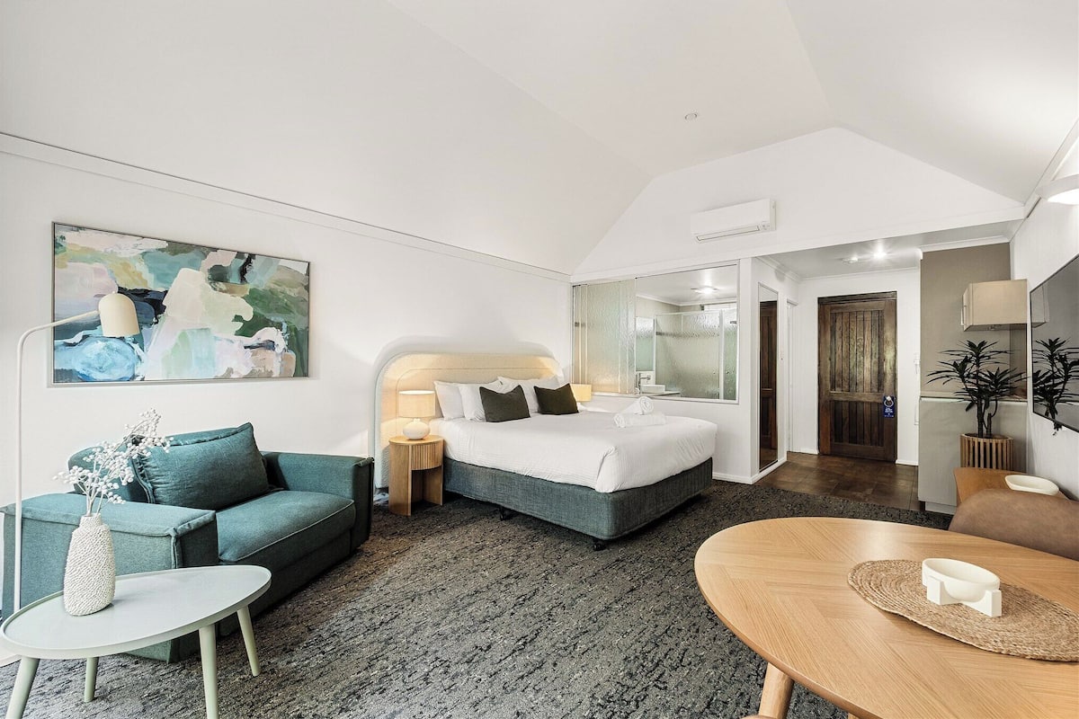 A modern hotel room with a bed, a sofa, a dining table, and a painting on the wall.