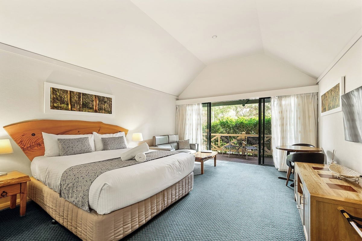 A hotel room with a large bed, a desk, a chair, and a balcony with a view of greenery.