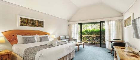 A hotel room with a large bed, a desk, a chair, and a balcony with a view of greenery.