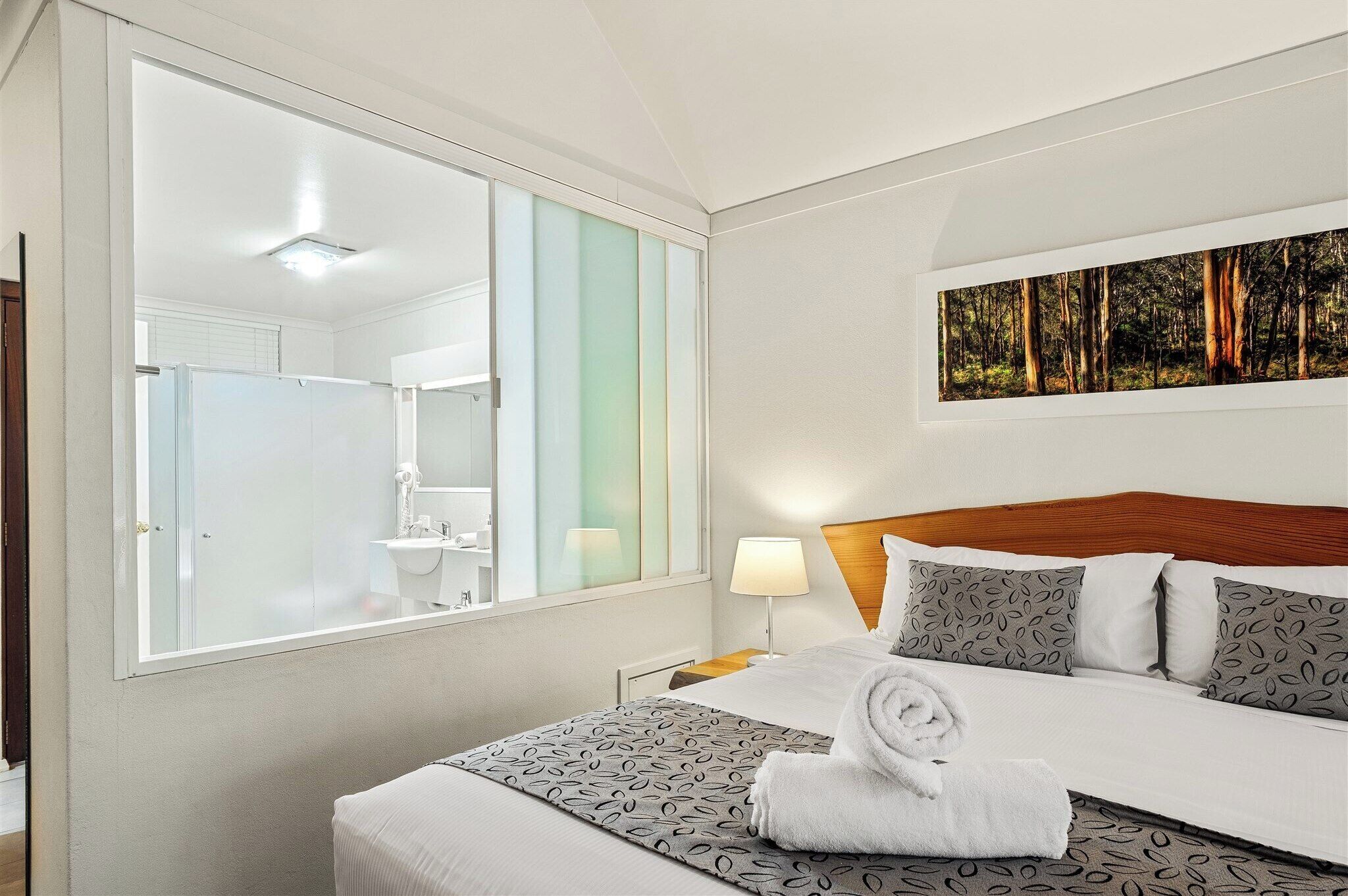 A hotel room with a large mirror, a bed with patterned bedding, a bedside lamp, and a painting of a forest on the wall.