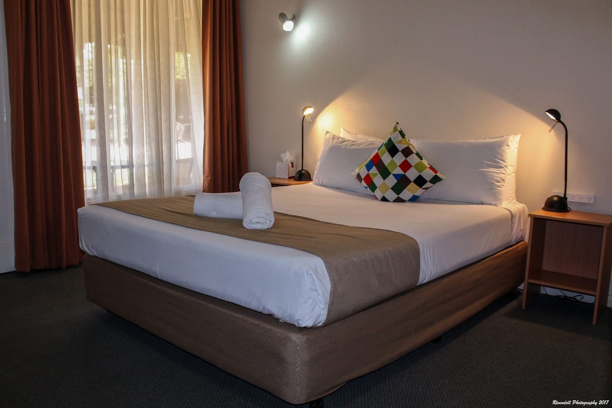 A neatly made bed with a brown headboard, white linens, and a colorful pillow. There is a nightstand with a lamp and a tissue box beside the bed.