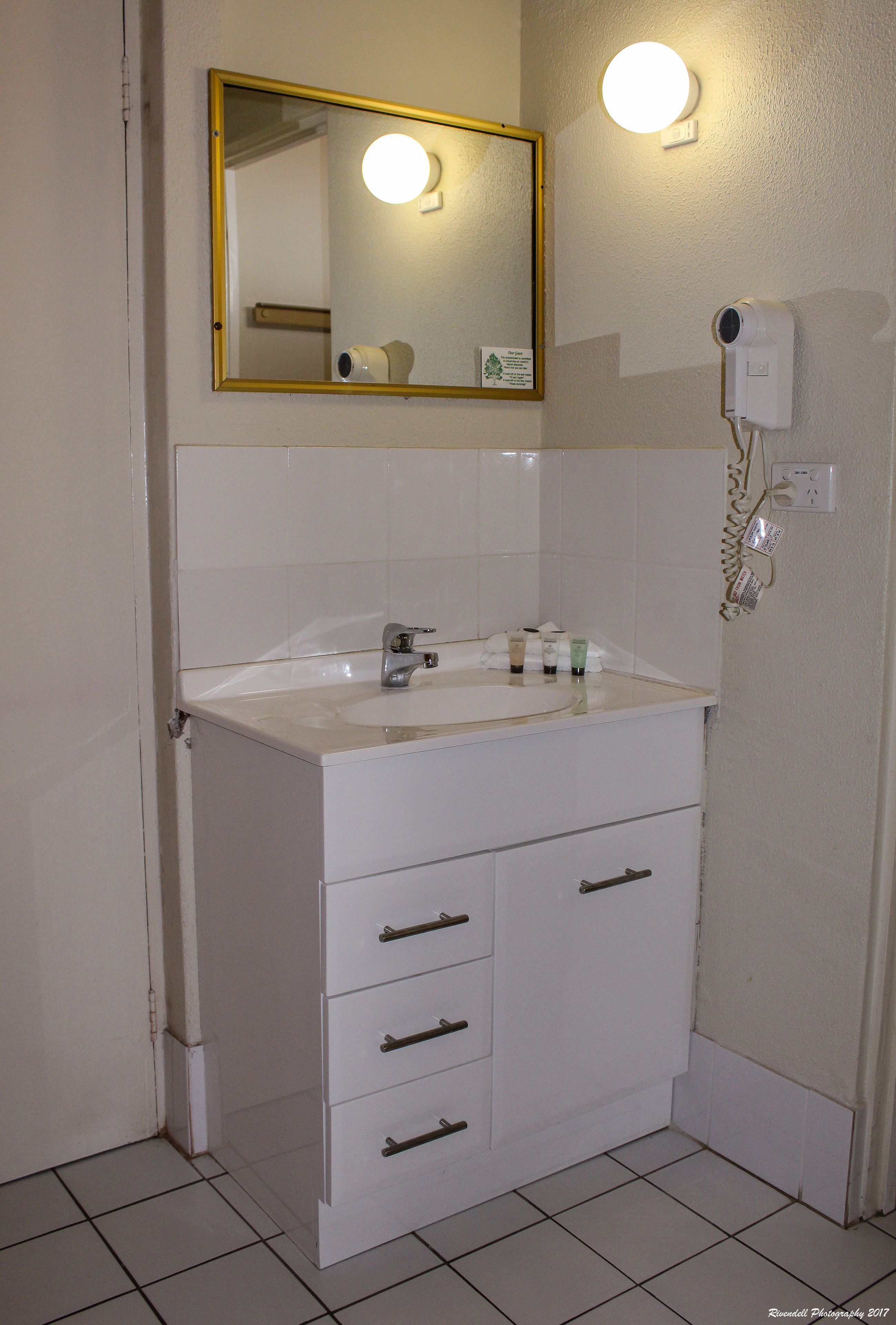 Casa de banho com lavatório branco, espelho e secador de cabelo na parede.