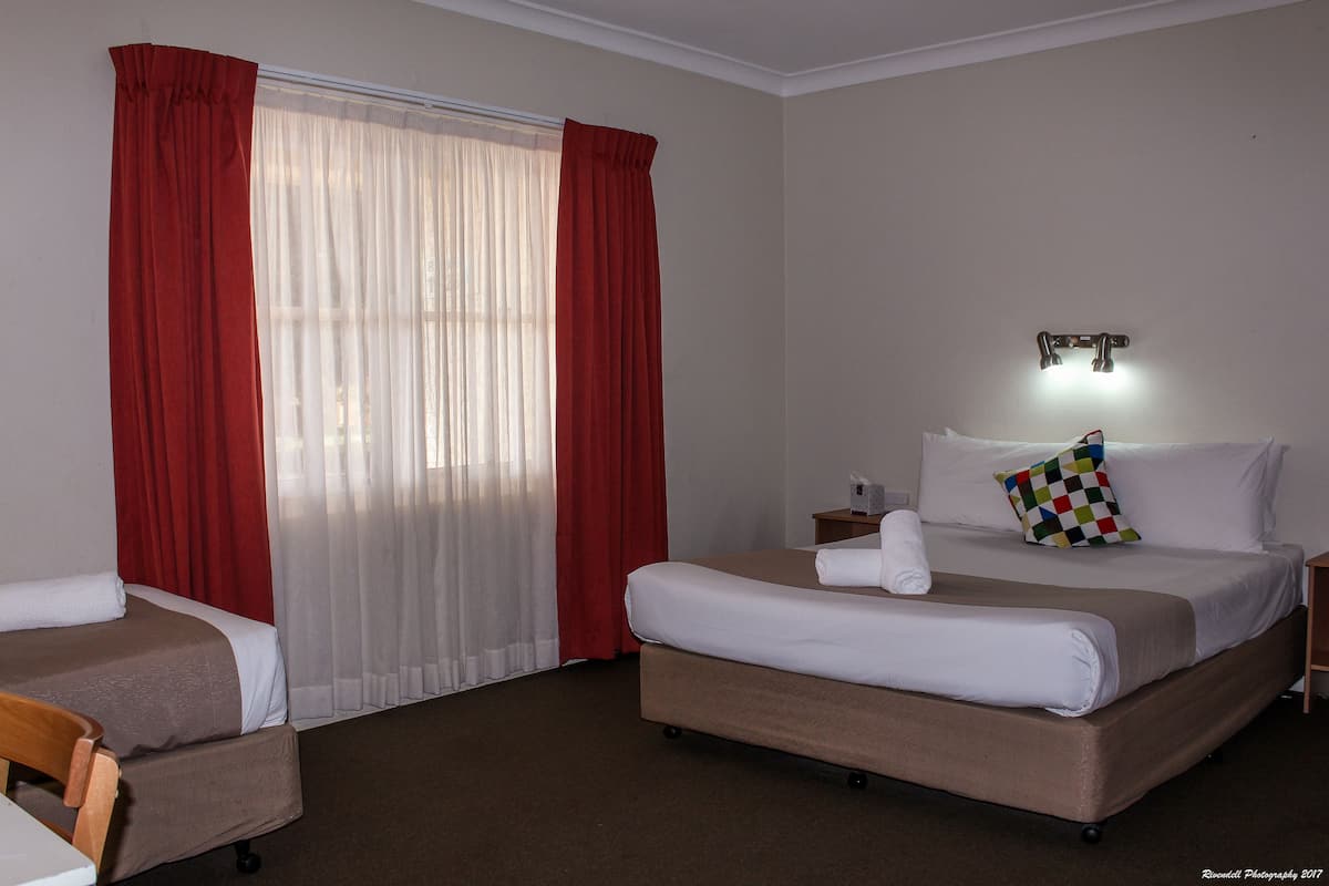 A hotel room with a bed, two bedside tables, a chair, a window with red curtains, and a wall-mounted light.