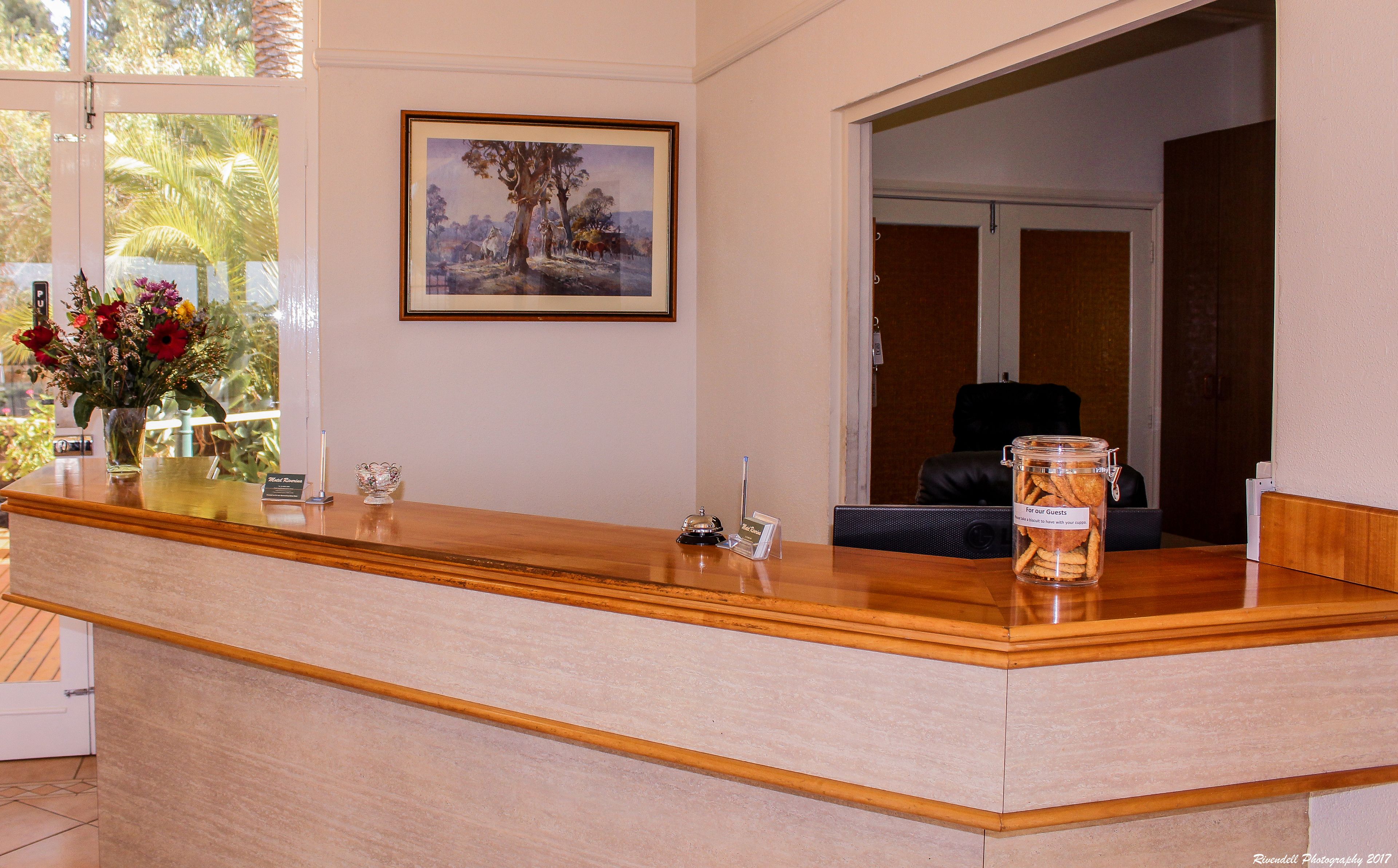 A hotel reception desk with a framed landscape painting, a vase of flowers, and a jar of snacks for guests.