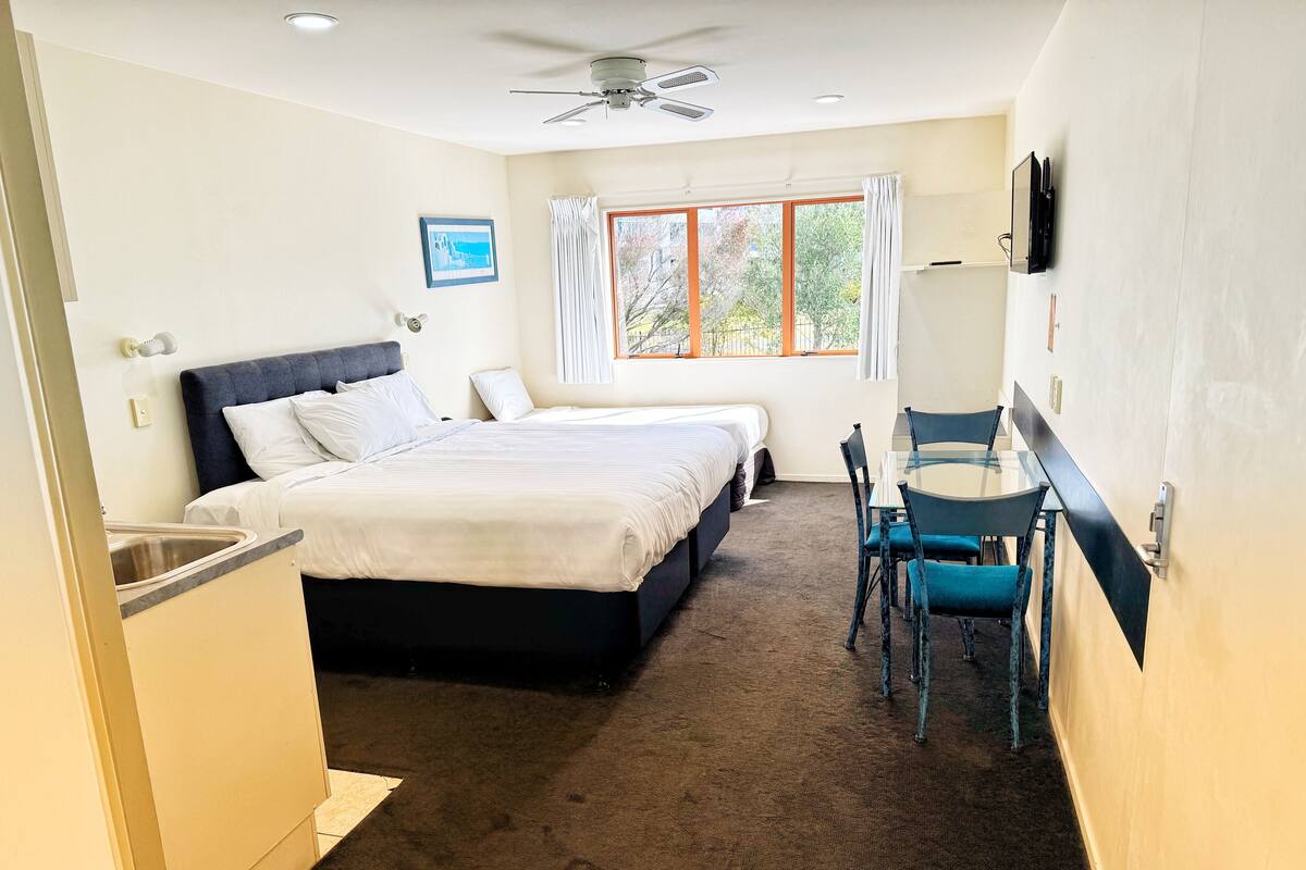 A hotel room with a bed, a desk, a chair, a window with curtains, a ceiling fan, and a TV mounted on the wall.