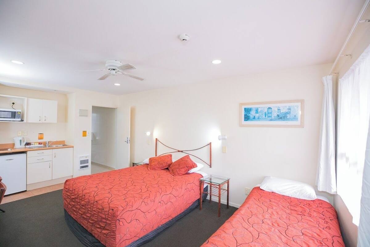 A hotel room with two beds, a kitchenette, a ceiling fan, and a painting on the wall.