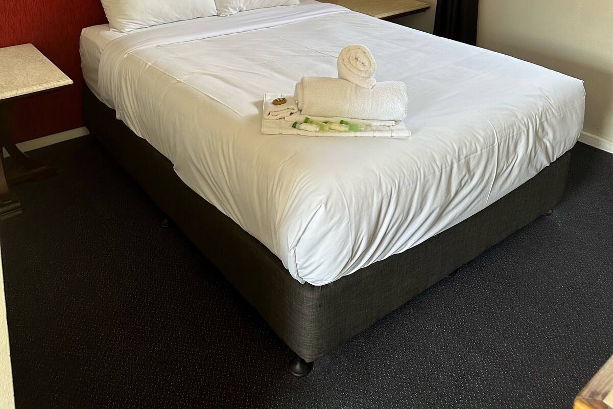 A hotel room with a neatly made bed, a red headboard, a bedside table with a lamp, and a window with curtains.