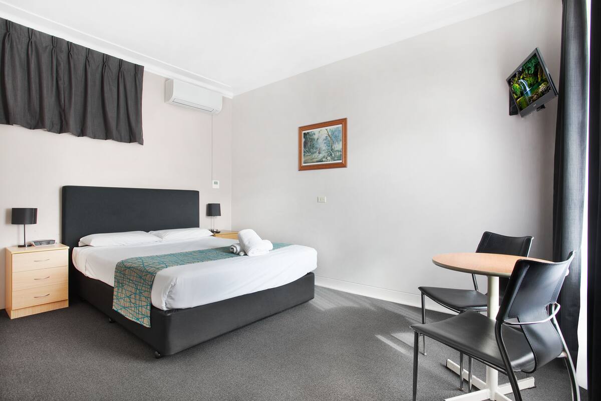 A hotel room with a bed, bedside tables, a desk with chairs, a painting on the wall, and a TV mounted on the wall.