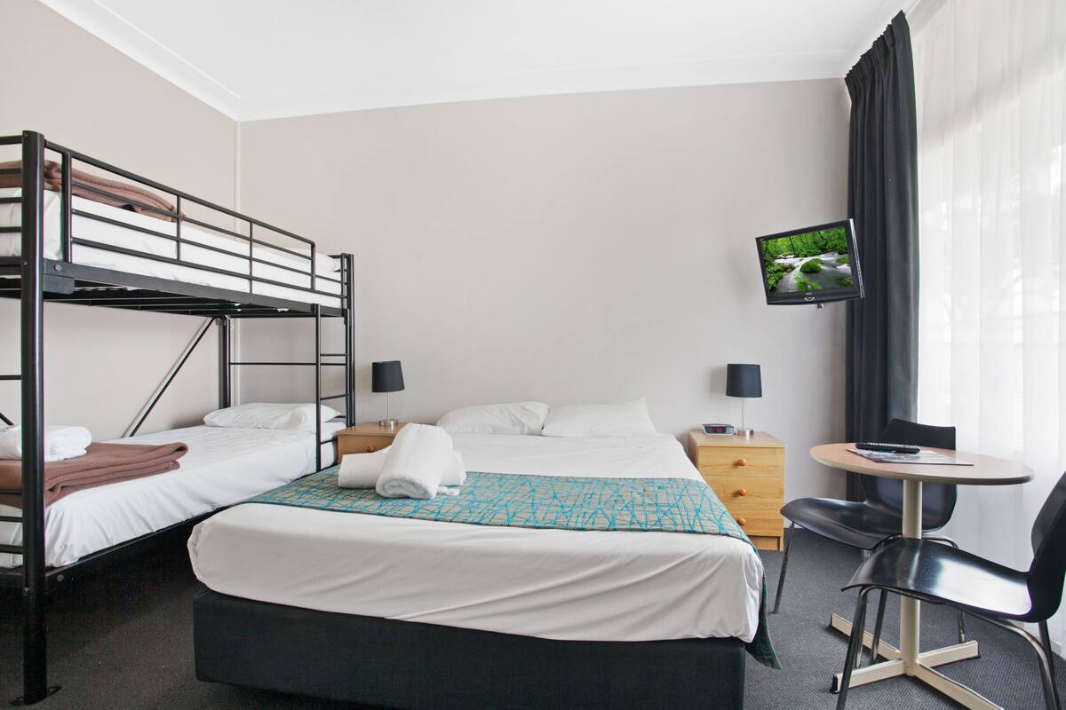 A hotel room with a bunk bed, a single bed, a TV, a desk with a chair, and a window with curtains.