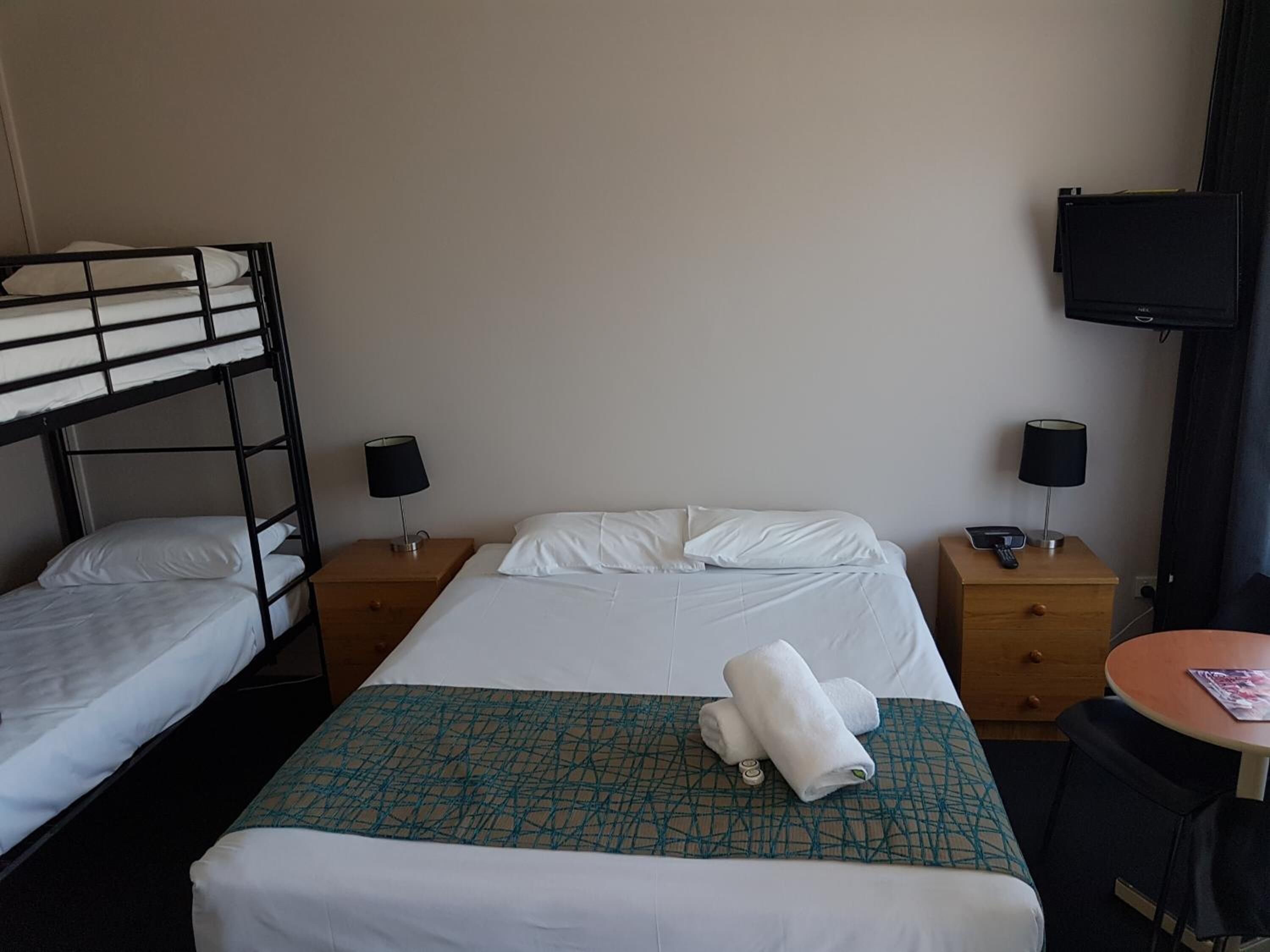 Een hotelkamer met een stapelbed, een enkel bed, twee nachtkastjes met lampen, een televisie aan de muur en een klein rond tafeltje met een tijdschrift.
