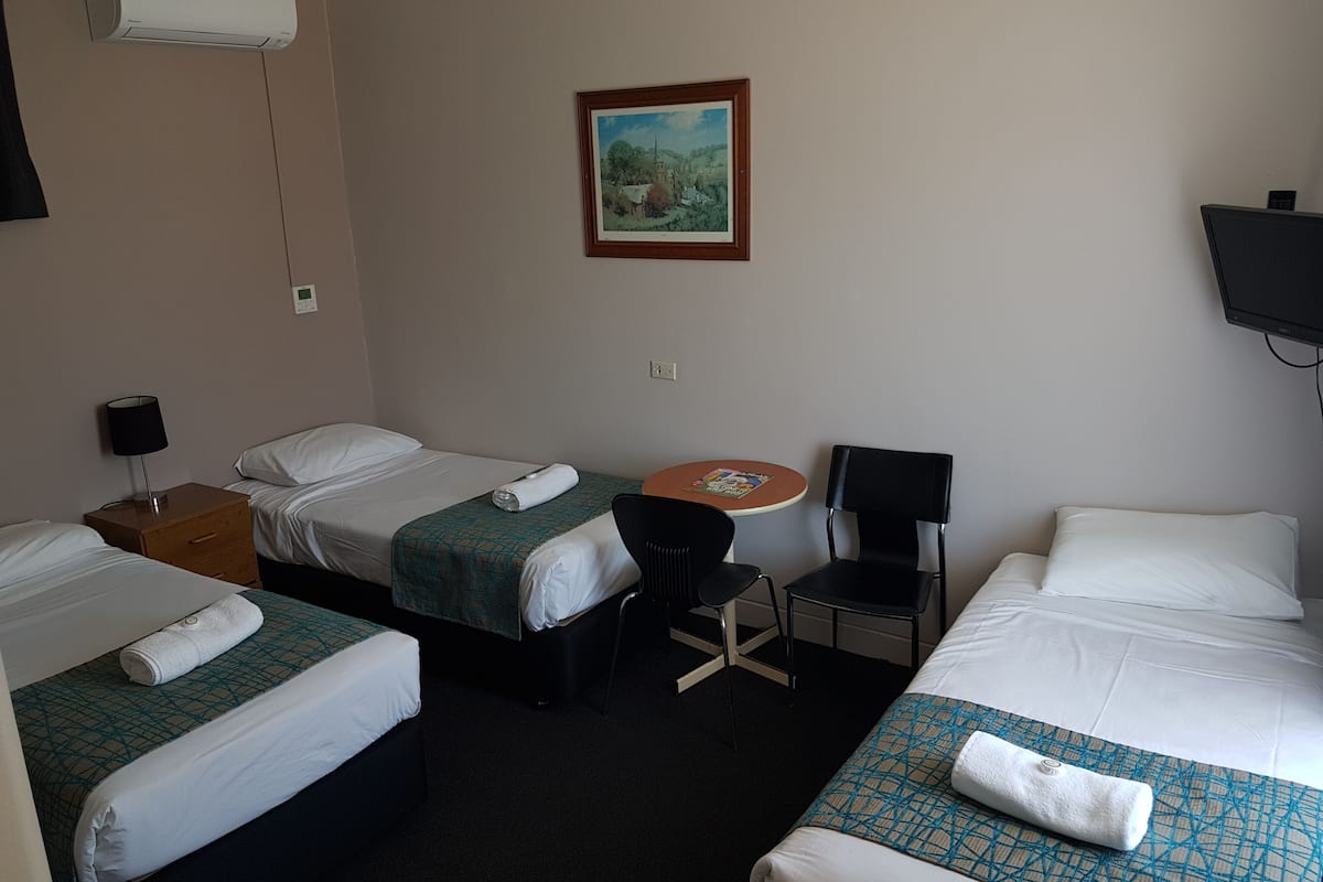 A hotel room with two single beds, a small table with a chair, a TV, and an air conditioning unit.