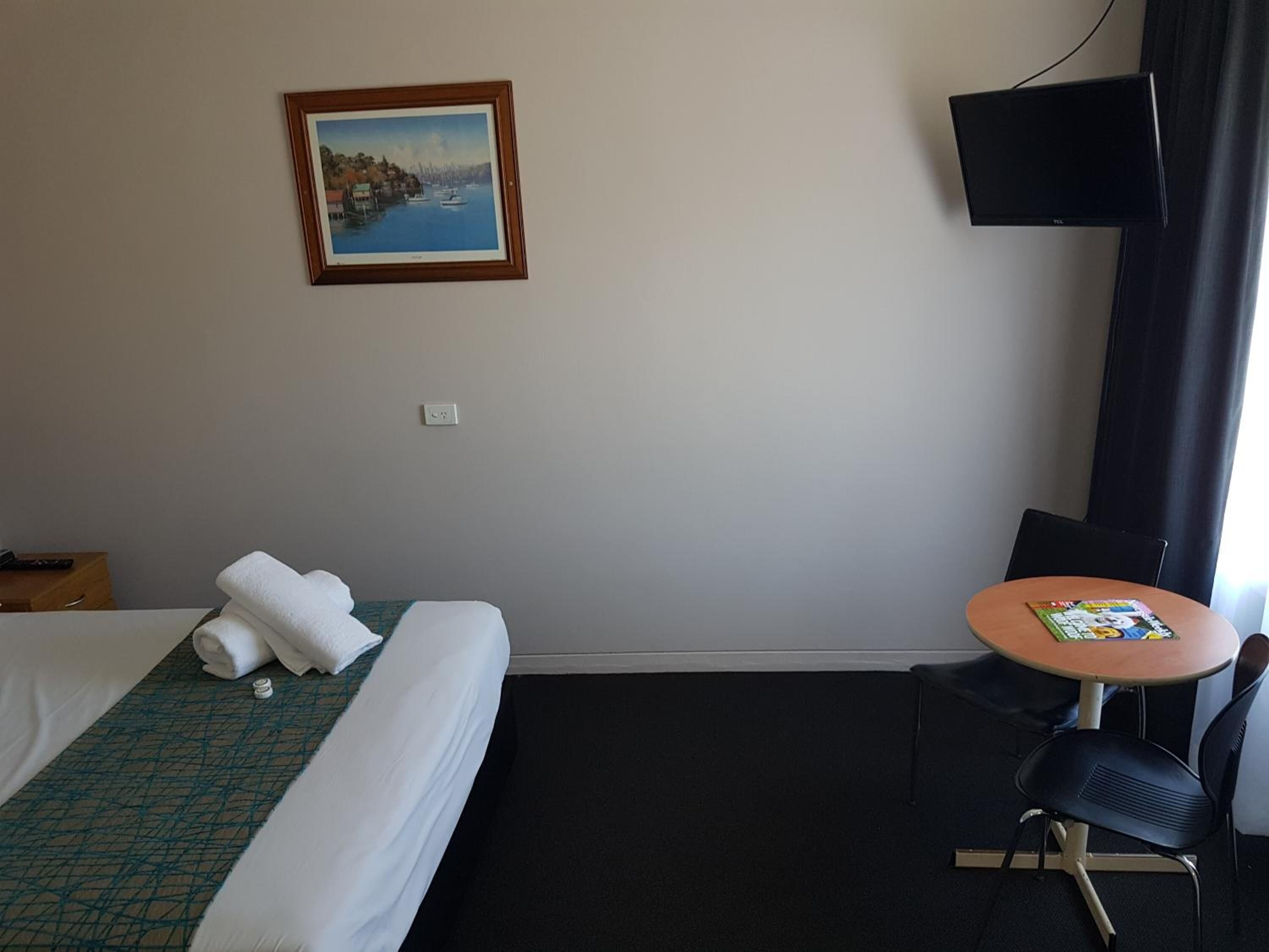 A hotel room with a bed, a small table with a chair, a painting on the wall, and a TV mounted on the wall.