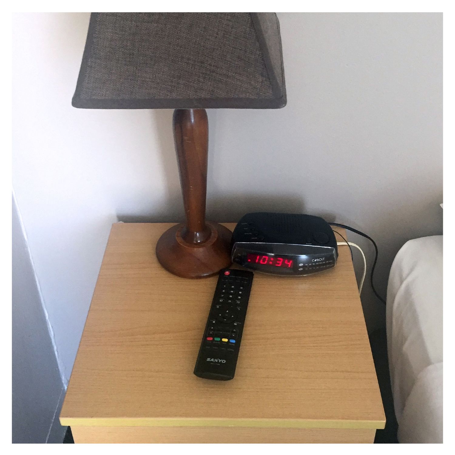 A wooden nightstand with a lamp, a digital clock, and a remote control.