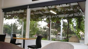A seating area with a table and chairs, large windows with a view of trees and a street, and a hanging plant. - Murray River Motel (Swan Hill)