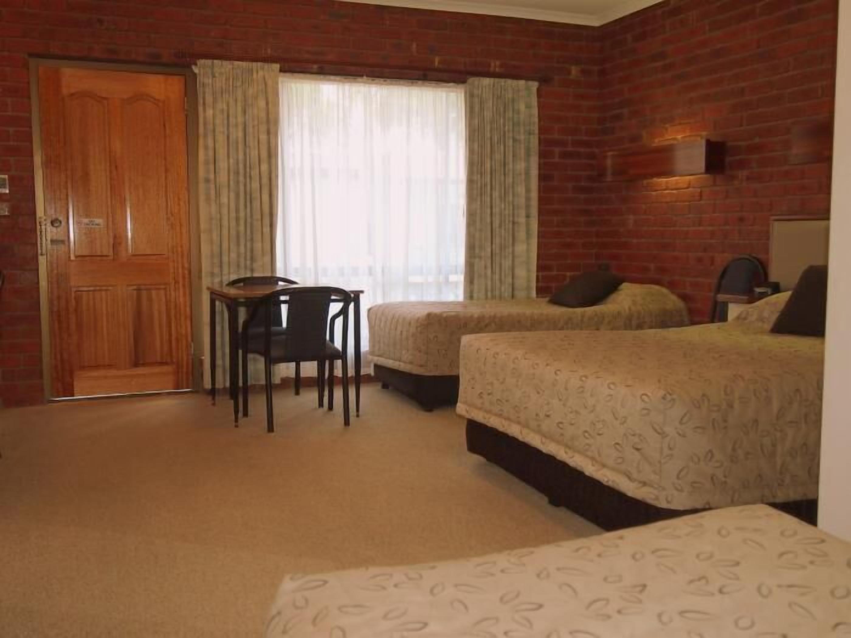 A hotel room with a brick wall, two beds, a wooden door, a small table with a chair, and a window with curtains.