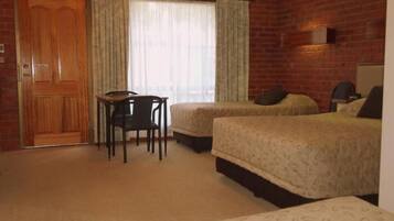 A hotel room with a brick wall, two beds, a wooden door, a small table with a chair, and a window with curtains.