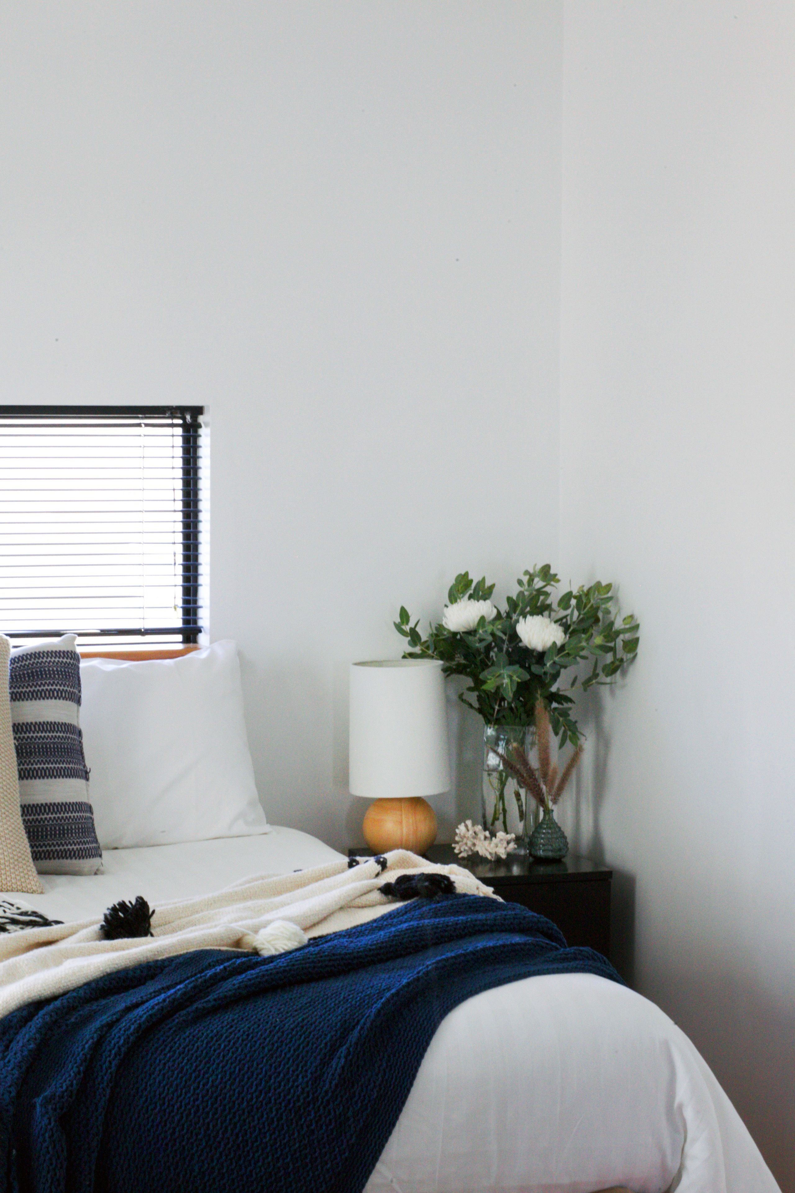 A neatly made bed with a blue blanket, white pillows, and a striped throw. A vase with white flowers and a lamp are on a bedside table.