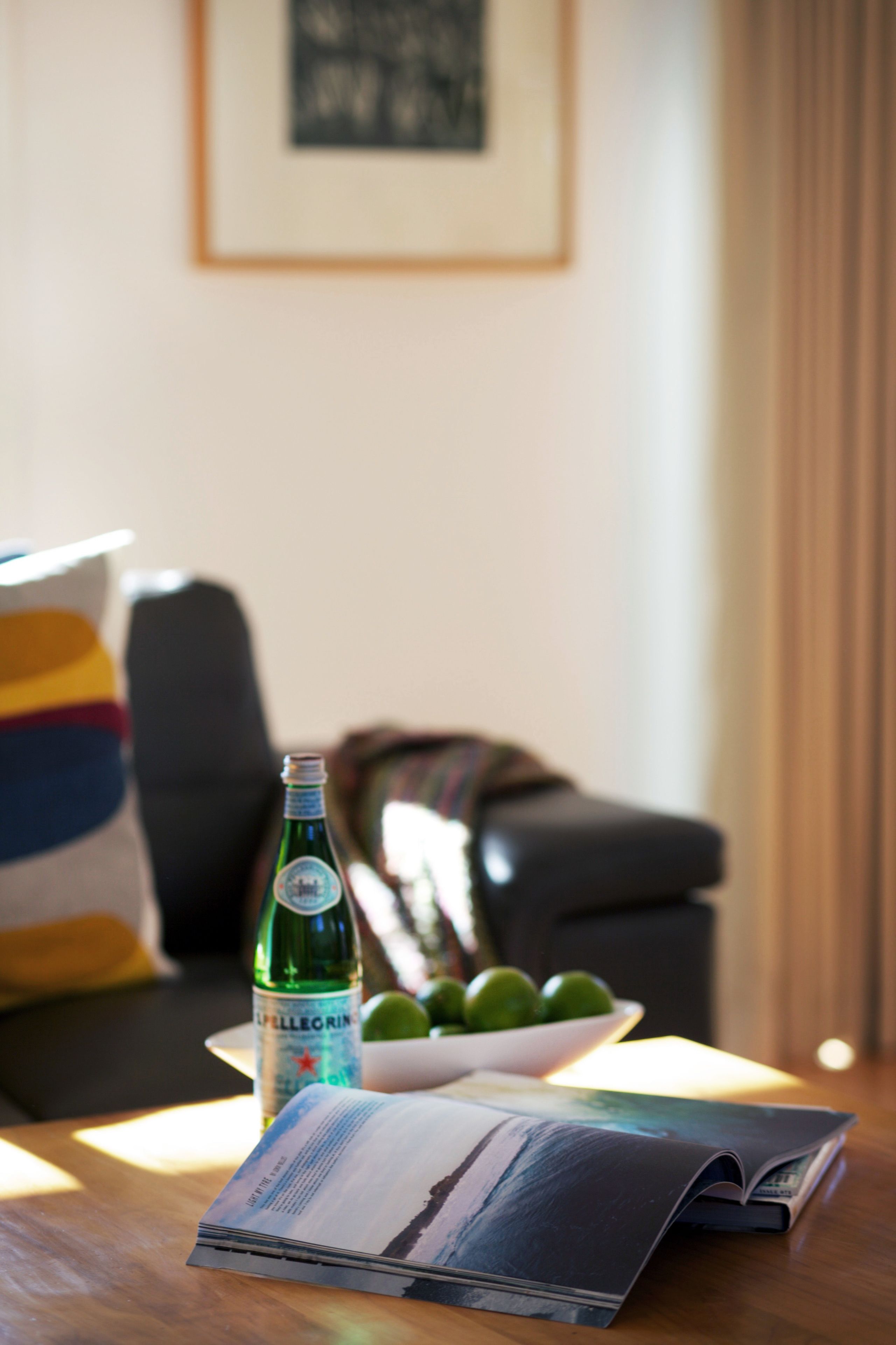 Une bouteille d’eau Pellegrino, un bol de citrons verts et un magazine sur une table en bois dans un salon, avec un canapé et un tableau encadré au mur.