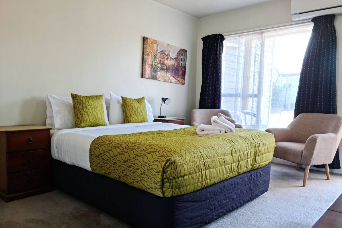 Una habitación de hotel con cama, una silla, una mesita de noche, una lámpara y un cuadro en la pared.