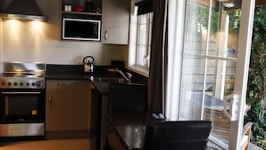 A compact kitchen with a stove, microwave, and sink. There's a window with curtains, and a chair is visible. - Cranford Cottages And Motel (Christchurch)