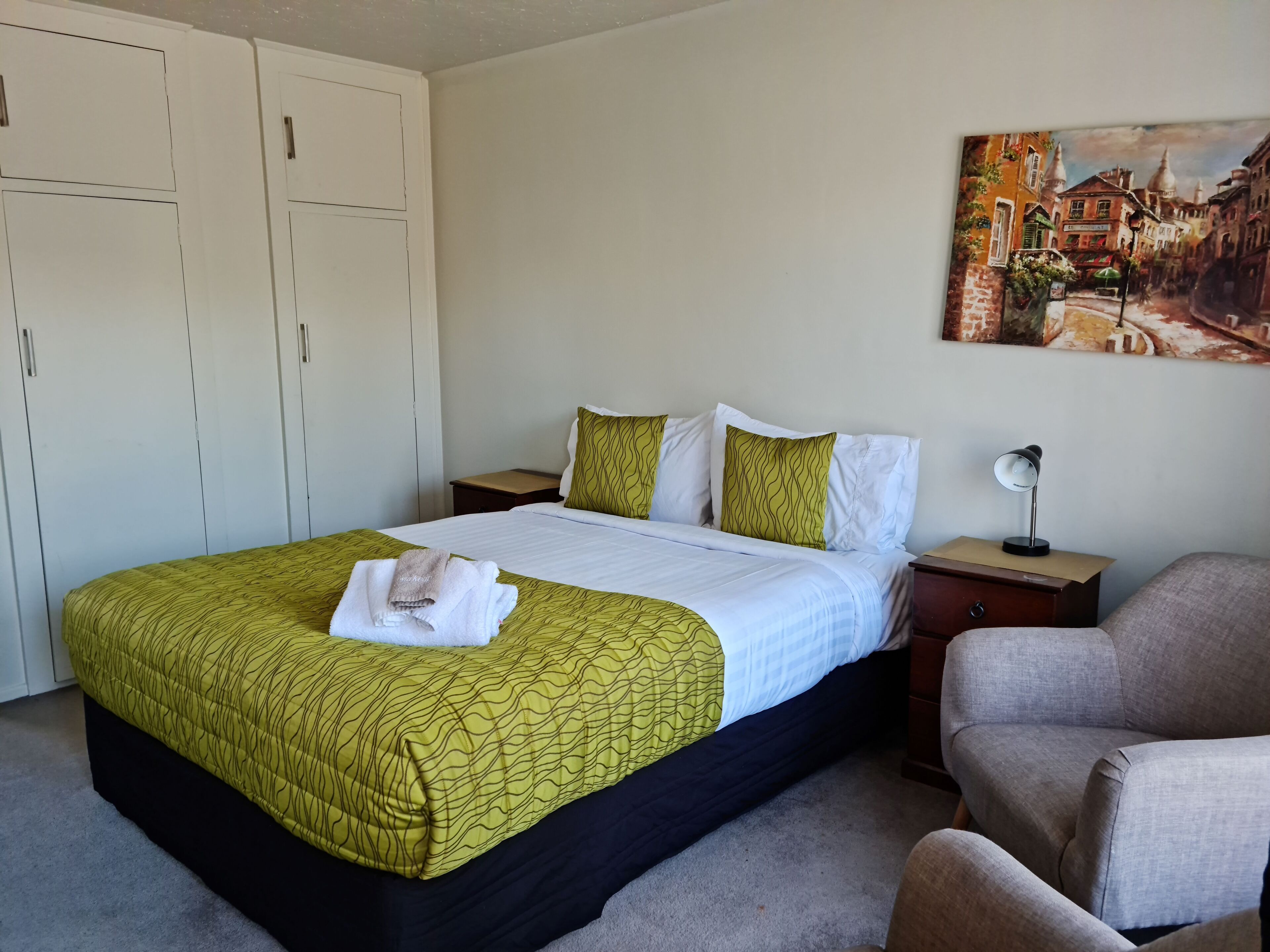 Une chambre d’hôtel avec un lit, une chaise, une table de chevet, une lampe et un tableau au mur.