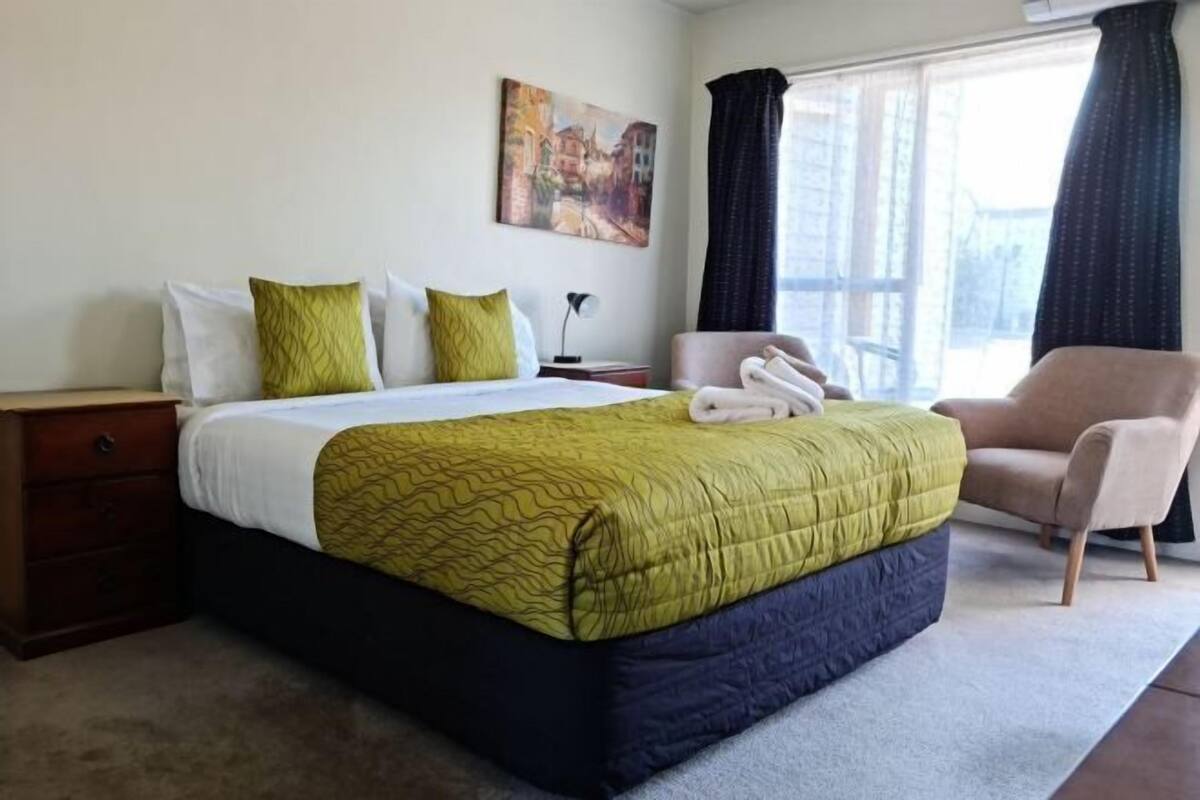 Una habitación de hotel con cama, una silla, una mesita de noche, un cuadro en la pared y una ventana con cortinas.