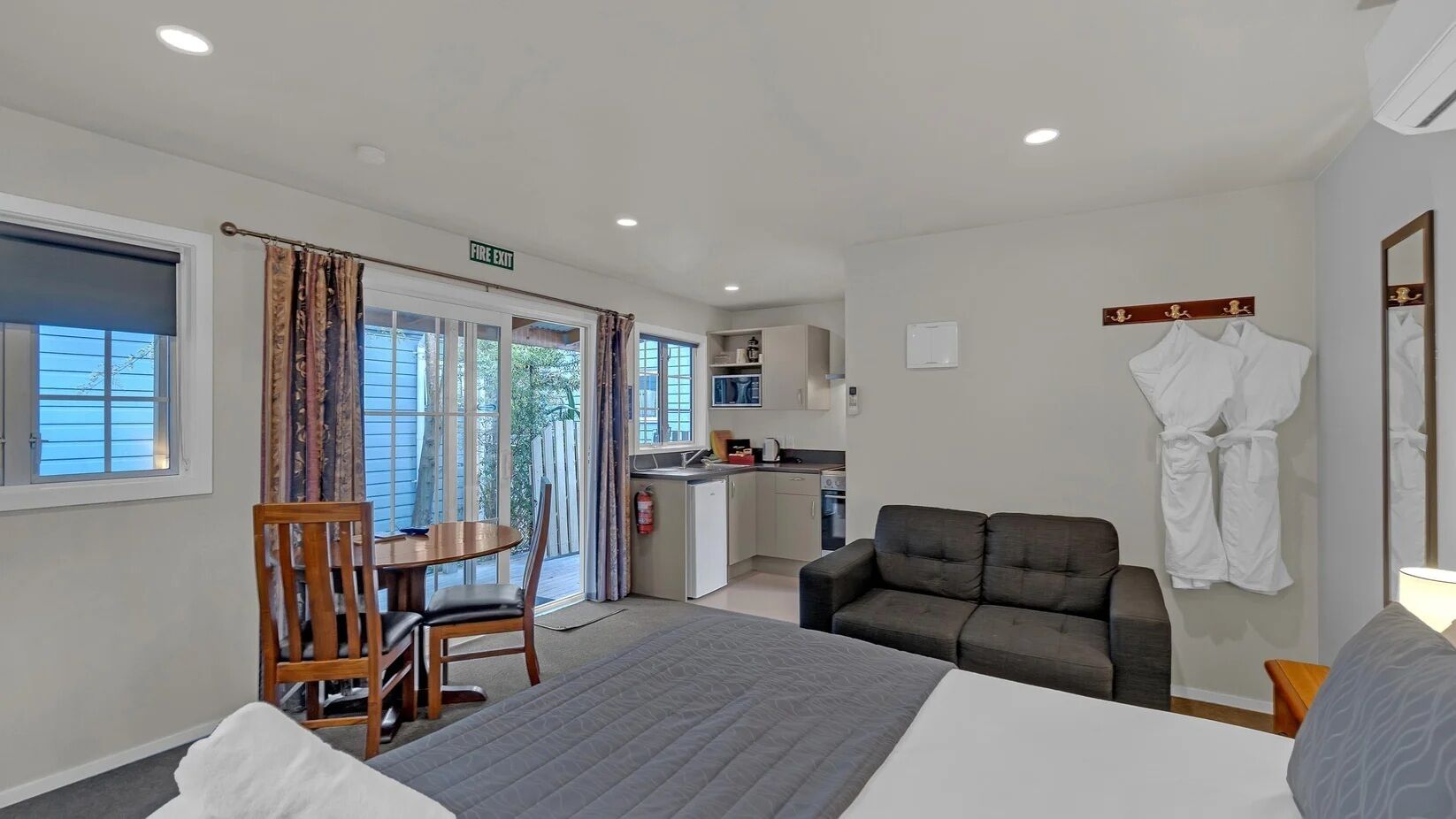 A hotel room with a bed, a sofa, a dining table with chairs, a kitchenette, and a fire exit sign.