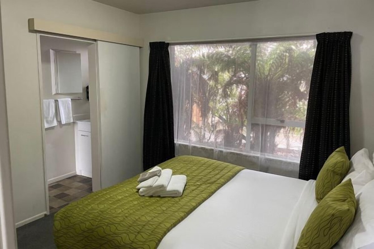 Habitación de hotel con cama, ventana con cortinas, armario y vistas a árboles.