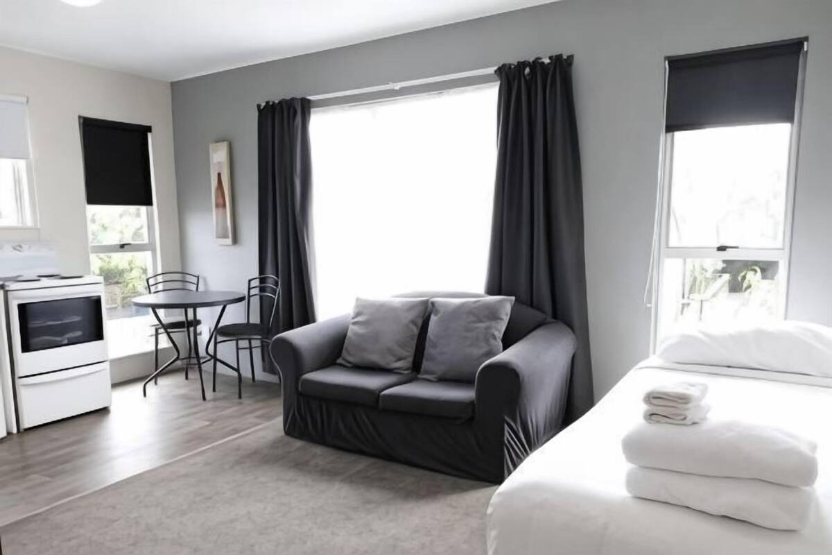 Una habitación moderna con sofá, una mesa de comedor, una cama con ropa blanca y un ventanal con cortinas.