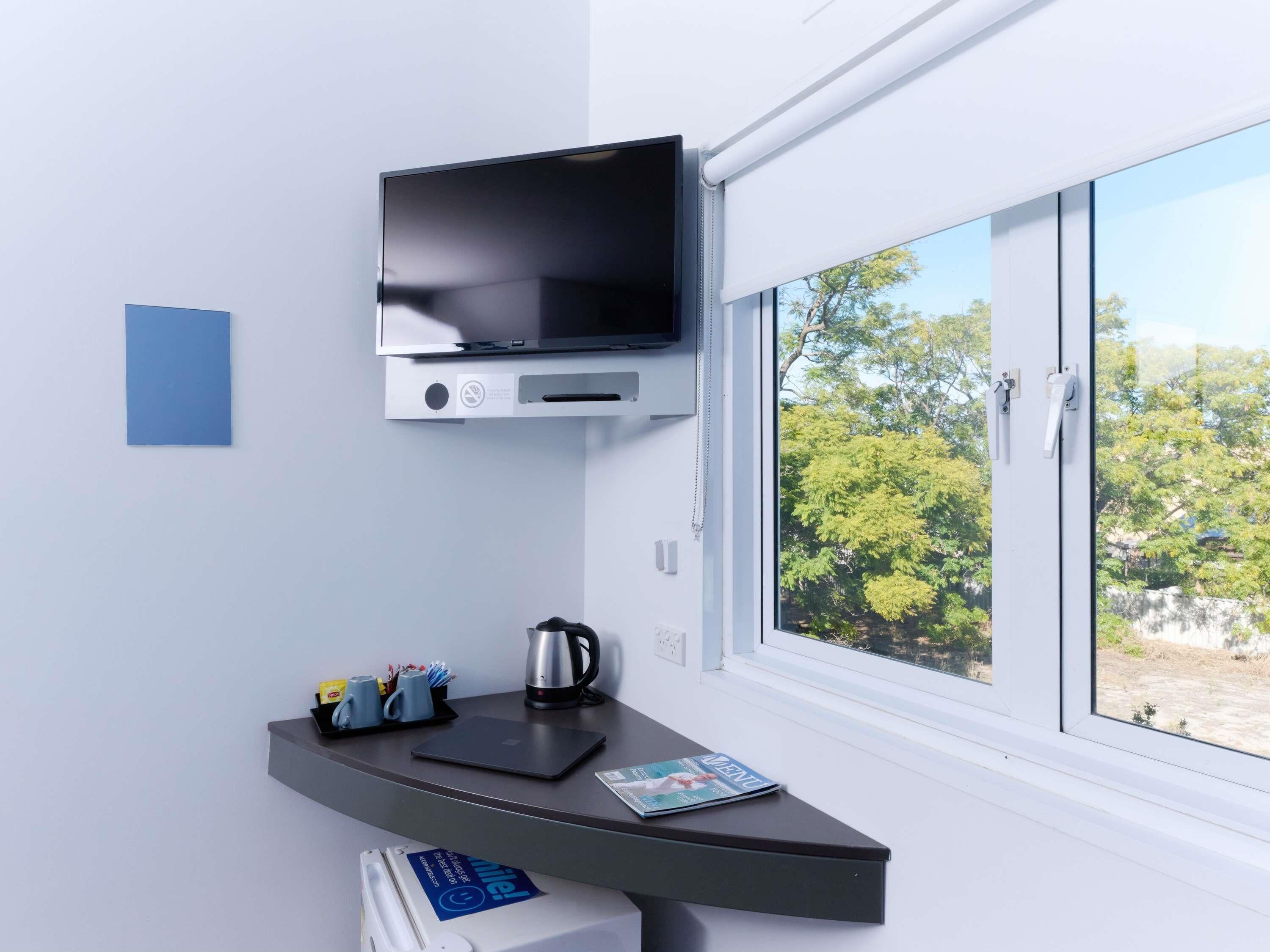 A modern room with a flat-screen TV mounted on the wall, a desk with a laptop, a kettle, and a magazine.