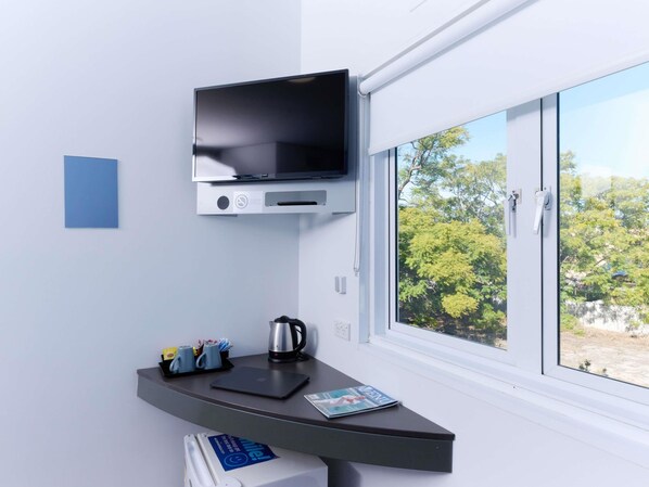 A modern room with a flat-screen TV mounted on the wall, a desk with a laptop, a kettle, and a magazine. - Ibis Budget Perth Airport (Redcliffe)