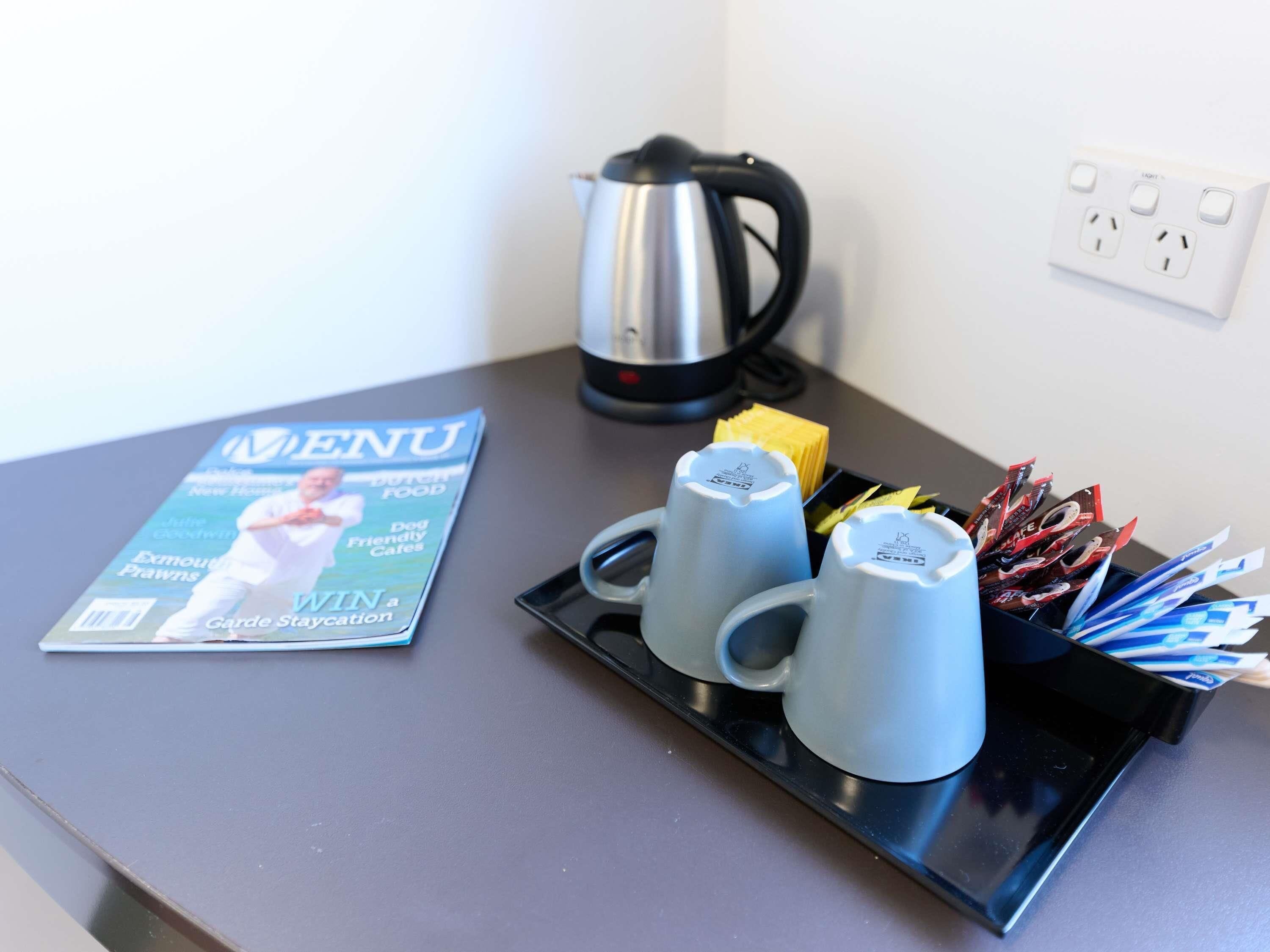 Una mesa con una revista, un hervidor, una bandeja con tazas y una caja de pajitas.