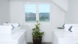 A laundry room with two washing machines, a potted plant, and a window with a view of a bridge and buildings. - Ibis Budget Perth Airport (Redcliffe)