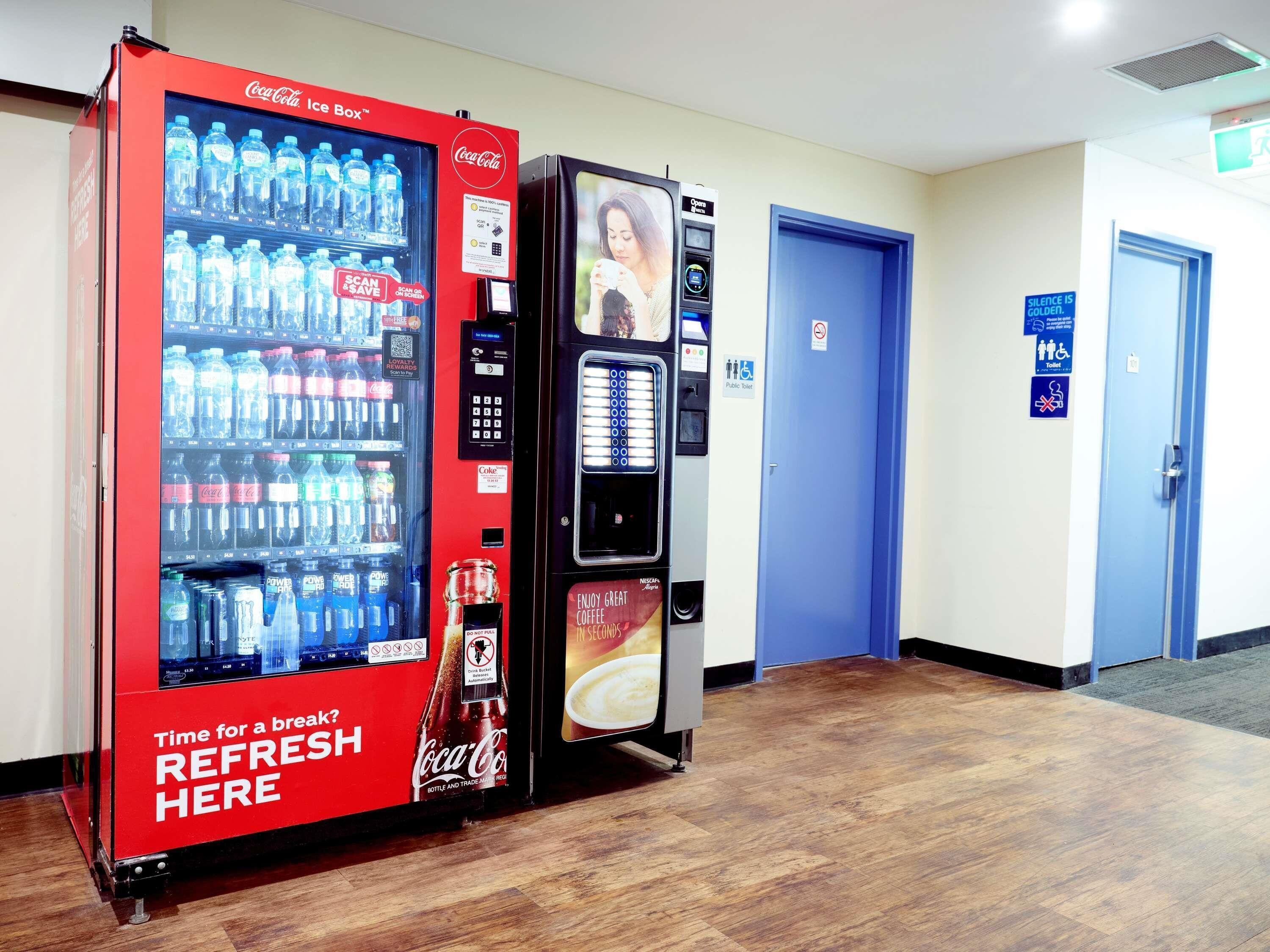 A vending machine area with Coca-Cola and Nescafé machines, a public toilet sign, and a blue door.