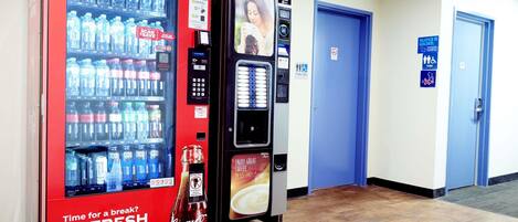 Área de máquinas de venda automática com máquinas de Coca-Cola e Nescafé, um sinal de banheiro público e uma porta azul.