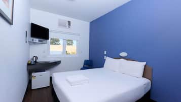 Quarto de hotel com cama, escrivaninha com computador, janela e televisão fixada na parede.