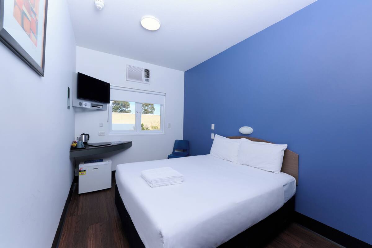 A hotel room with a bed, a desk with a flat-screen TV, a window, and a small kitchenette.