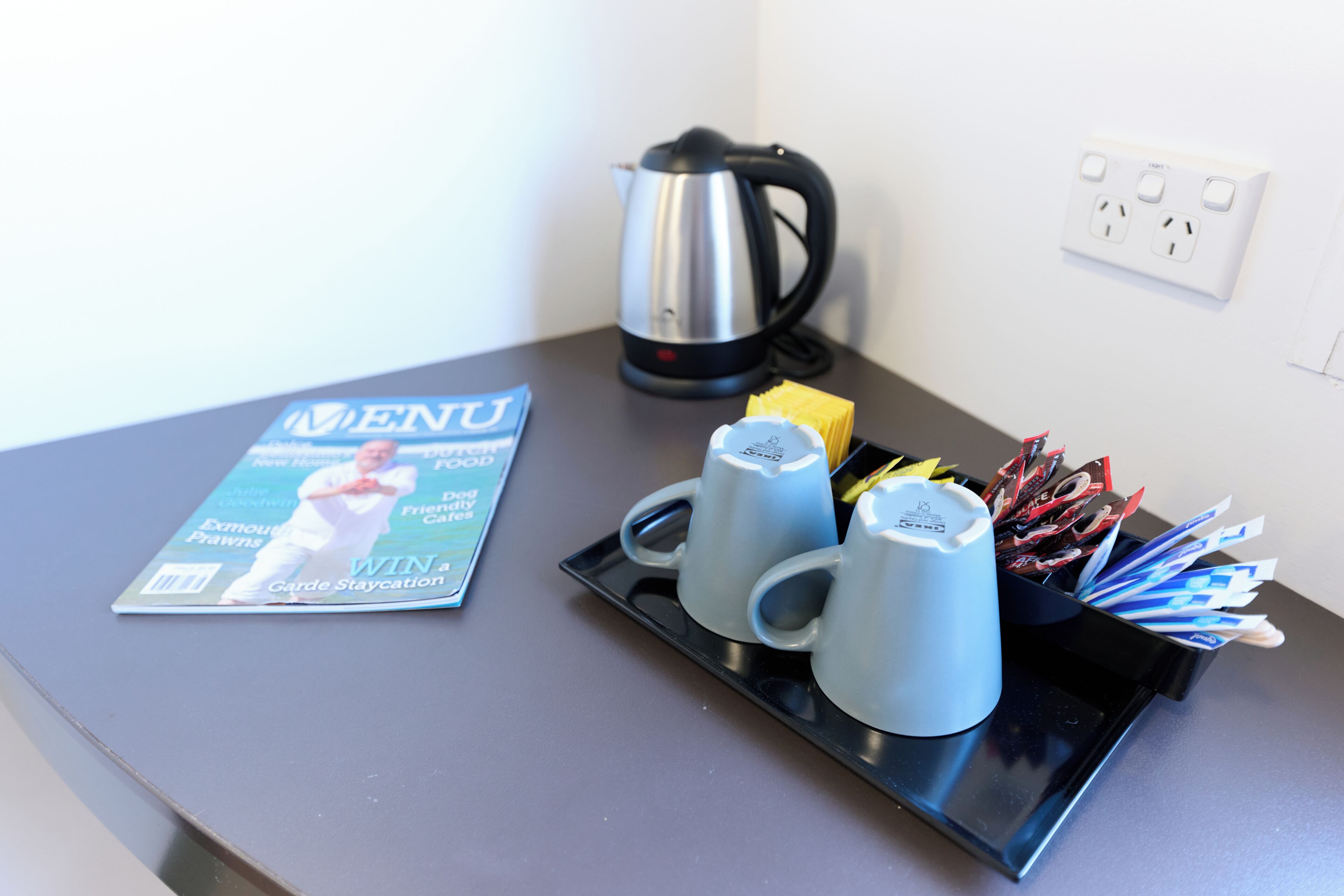 A table with a magazine, a kettle, and a tray with cups and utensils.