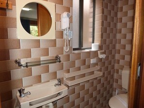 Un baño con inodoro, lavamanos y espejo. Hay un secador de pelo y un teléfono fijado a la pared.
